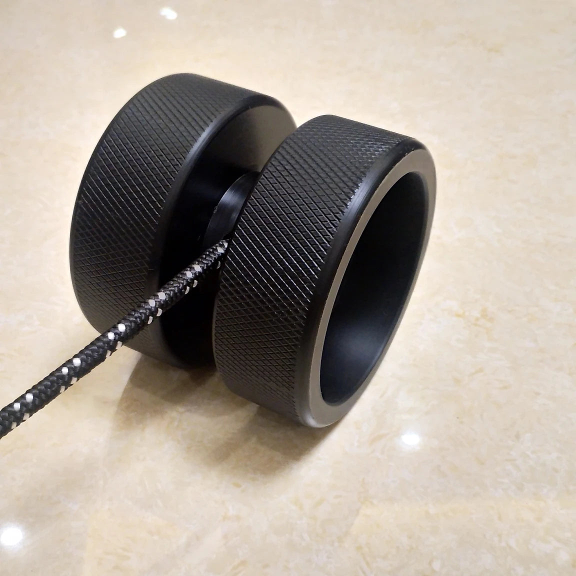 A close-up of the black Finger Grip and Forearm Strength Trainer is shown against a beige marble background. It features a textured surface and a black-and-white string extending to the left.
