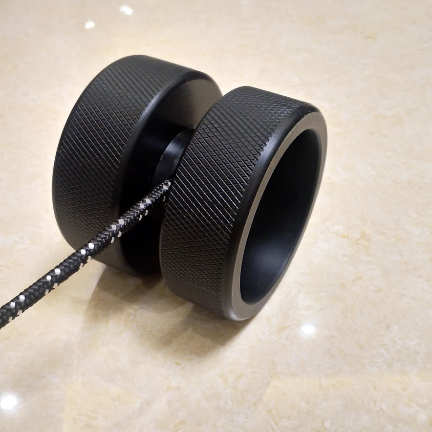 A close-up of the black Finger Grip and Forearm Strength Trainer is shown against a beige marble background. It features a textured surface and a black-and-white string extending to the left.