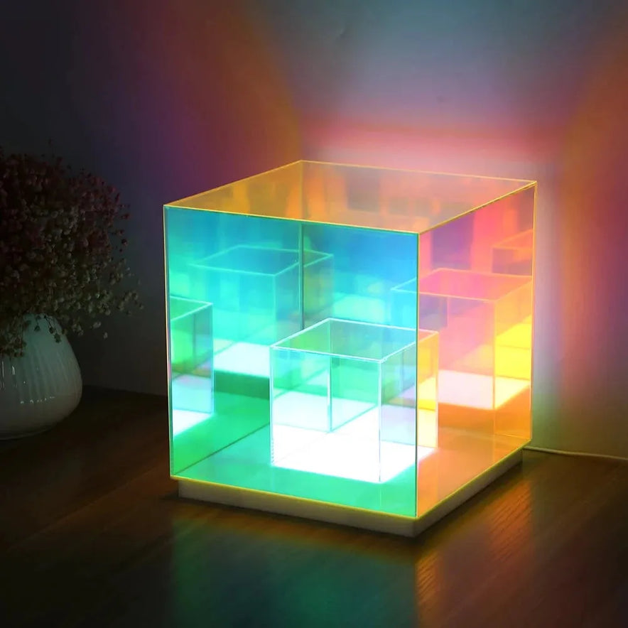 The Vibrant RGB LED Night Light features a glowing cube design with smaller translucent cubes inside, displaying dynamic blue, green, and orange colors. Ideal for parties or home décor, it excels as a versatile table lamp placed on wood beside flowers in soft lighting.