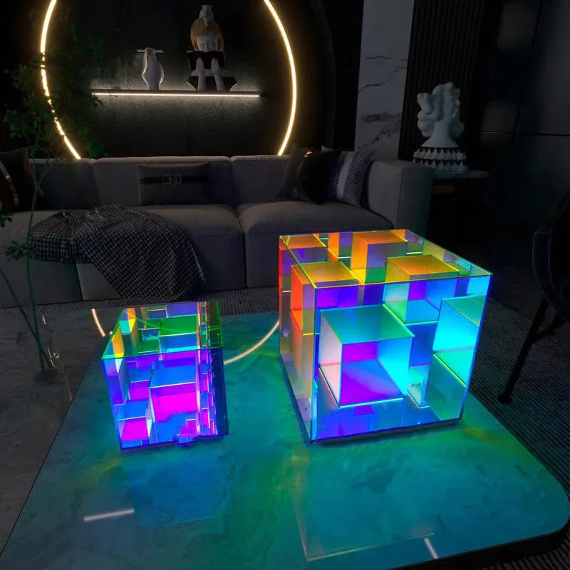 Two geometric, multicolored cubes from the Vibrant RGB LED Night Light for Parties and Home Décor sit on a reflective surface in a dimly lit room. The larger cube is behind the smaller, enhancing dynamic lighting. A couch and sculptures are visible with a circular wall light in the background.