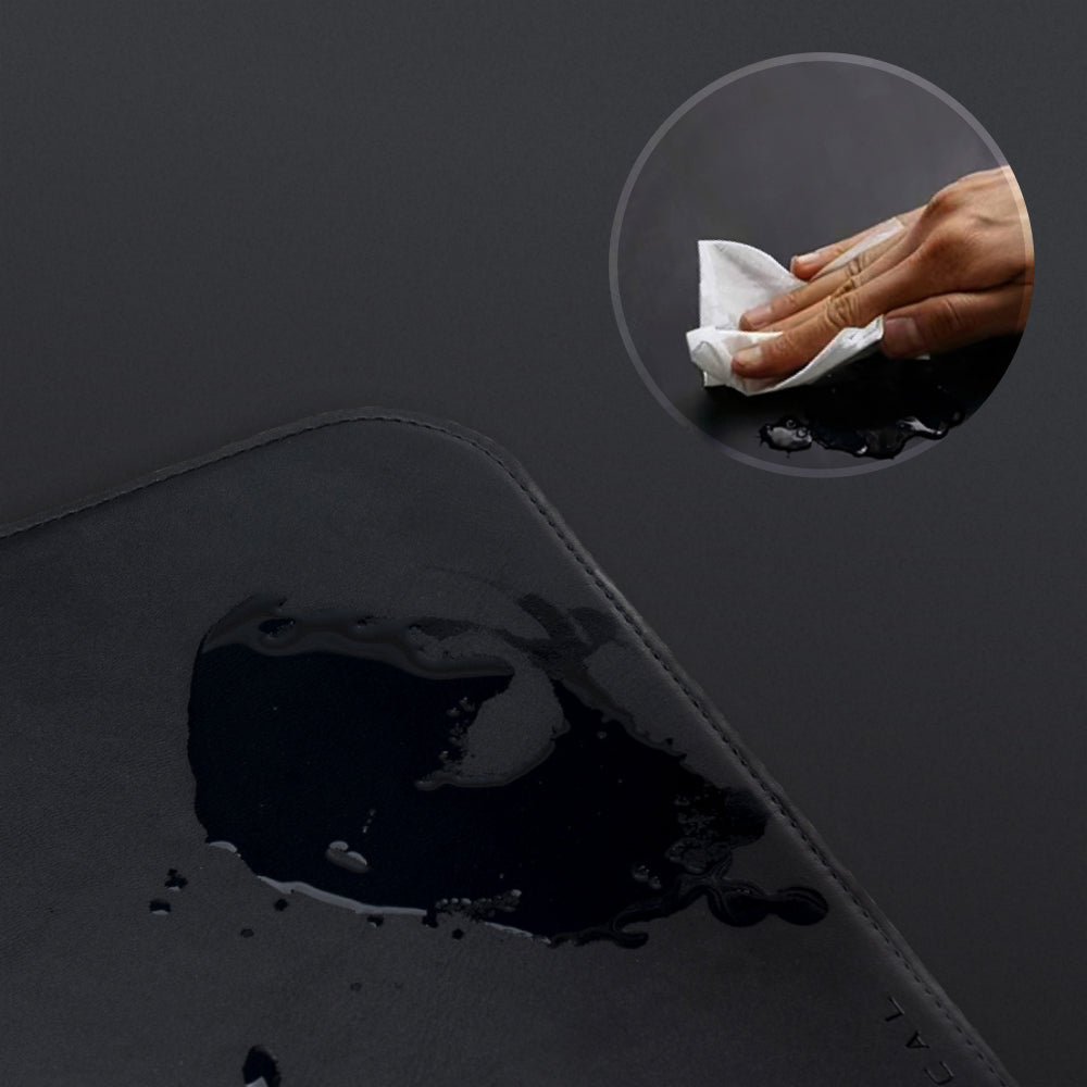 A close-up of the black Desk Mat Bundle with water spilled on its vegan leather surface. An inset shows a hand wiping it clean with a tissue, demonstrating the mat's easy-to-clean feature.