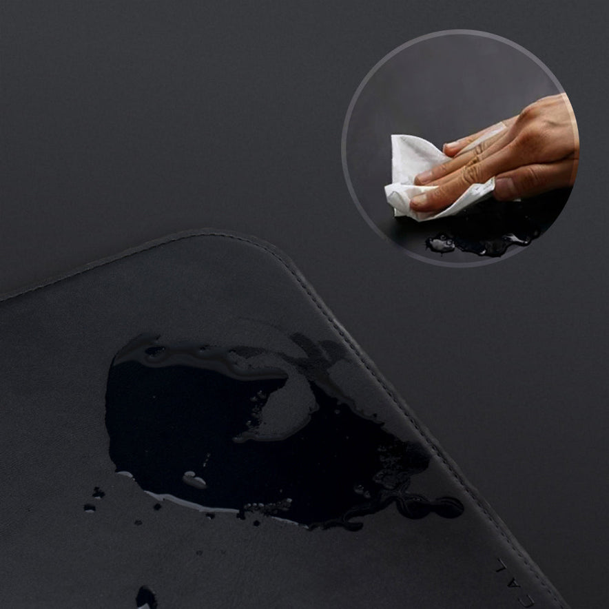 A close-up of the black Desk Mat Bundle with water spilled on its vegan leather surface. An inset shows a hand wiping it clean with a tissue, demonstrating the mat's easy-to-clean feature.