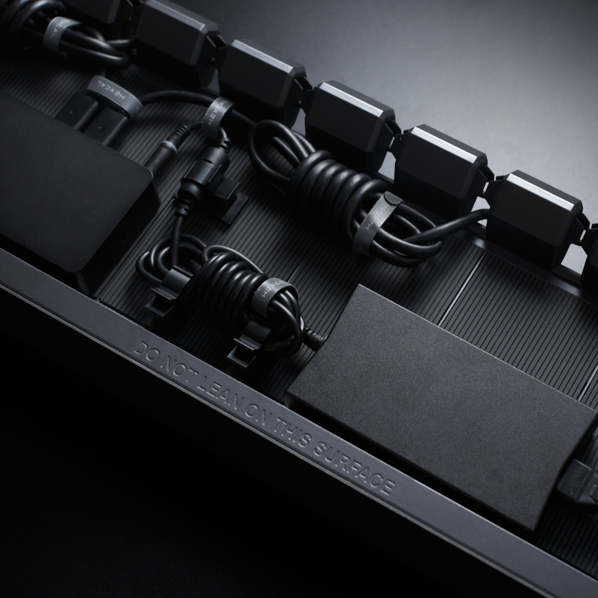 Close-up of Hexcal Studio™ shows a row of black electronic components and coiled cables with organized cable management on a dark surface, featuring "DO NOT LEAN ON THIS SURFACE" embossed near the bottom edge.
