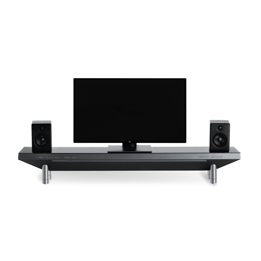 The Hexcal Studio™ is a modern home entertainment setup with a flat-screen TV, black speakers, and a sleek soundbar shelf with metallic legs and built-in cable management, all shown against a plain white background.