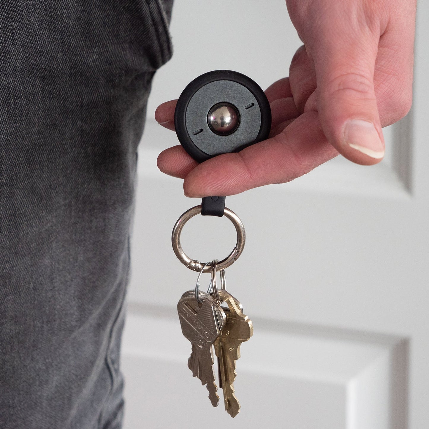 A hand holds the ONO Scroller Keychain—a black magnetic key holder with a metal ring, keychain attachment, and several keys—while wearing dark pants and standing before a white door.