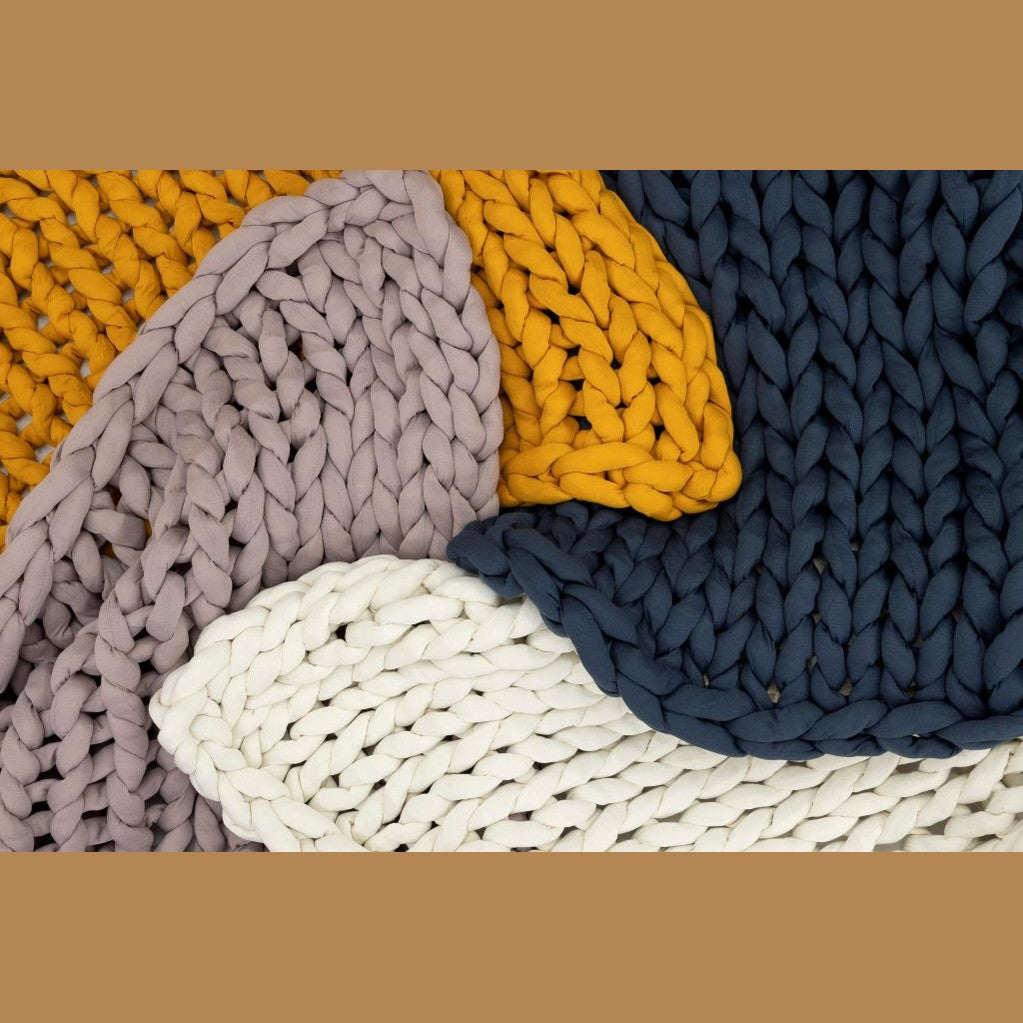 Close-up of chunky Knit Weighted Blankets in mustard yellow, navy blue, lavender, and cream—layered for a textured, cozy look. Enjoy the comfort and calming effect of a super soft weighted blanket with this inviting display.
