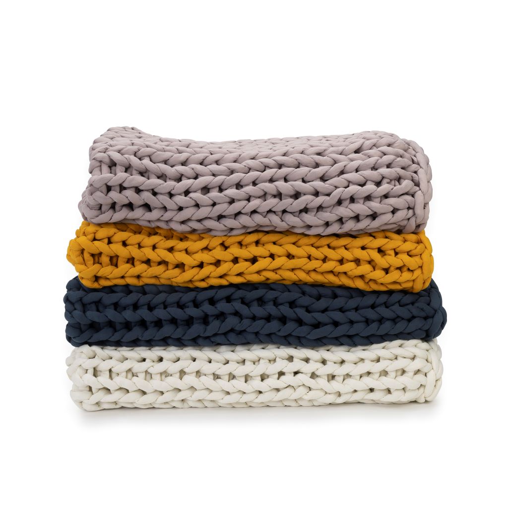 A stack of four Knit Weighted Blankets—super soft in light gray on top, then mustard yellow, dark navy blue, and cream below—all neatly folded against a white background.