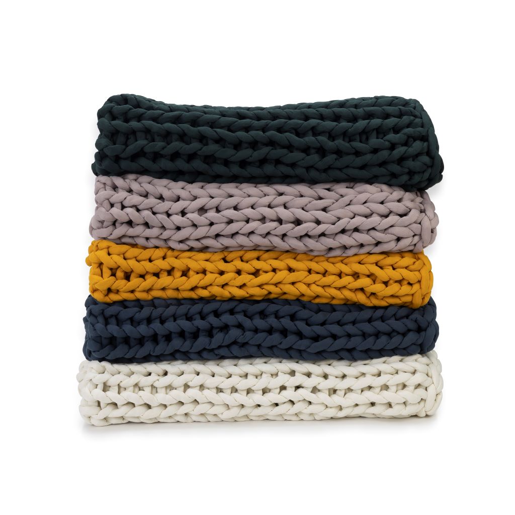 A stack of five Knit Weighted Blankets in dark green, light mauve, mustard yellow, navy blue, and white are neatly folded against a white background.