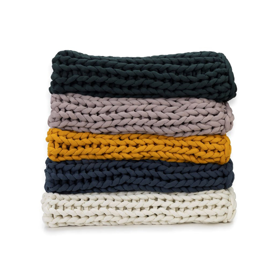 A stack of five Knit Weighted Blankets in dark green, light mauve, mustard yellow, navy blue, and white are neatly folded against a white background.