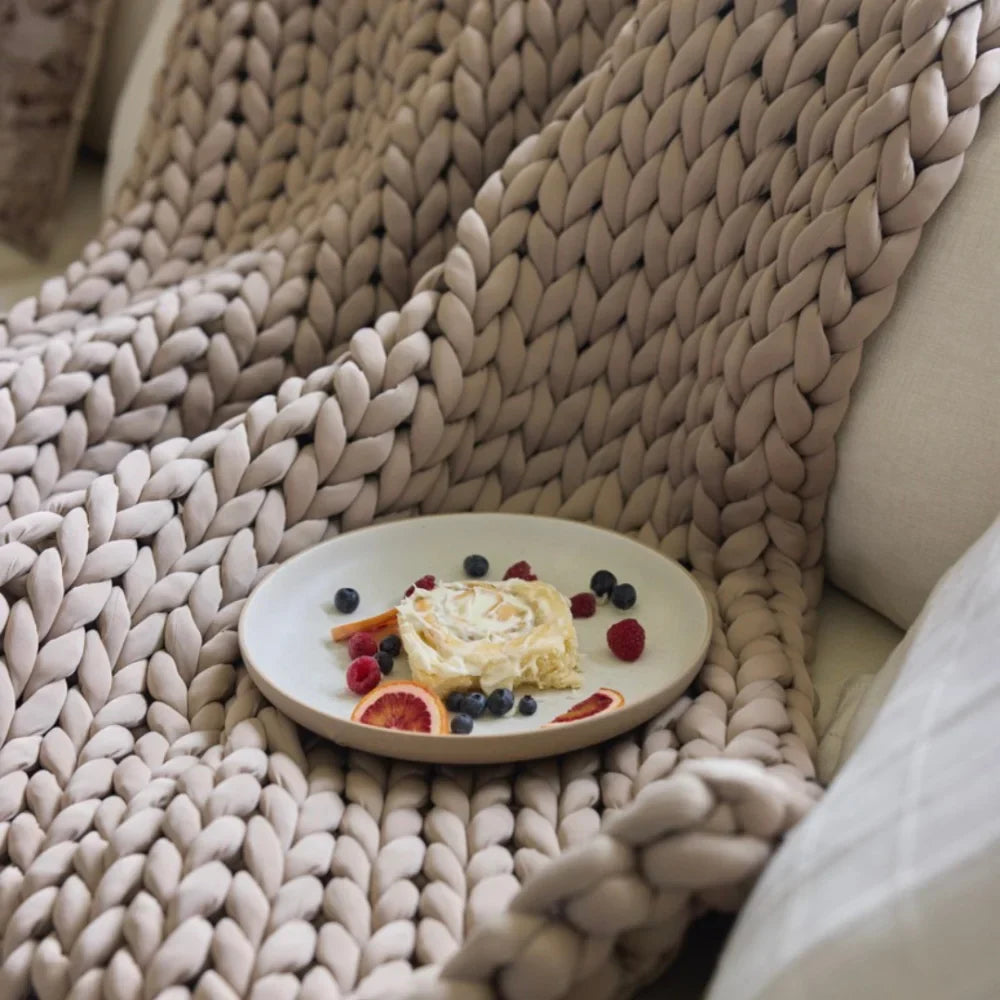 A plate with a creamy dessert, berries, and orange slices rests on the Ice Knit Weighted Blanket in Coastal Sand, which is draped over a light-colored sofa.