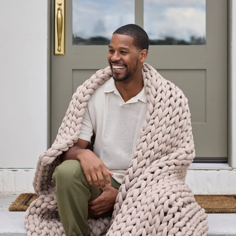A smiling man sits on a doorstep wrapped in the Ice Knit Weighted Blanket, a chunky beige cooling blanket. He wears a light short-sleeve shirt and olive green pants, with a door and welcome mat behind him.