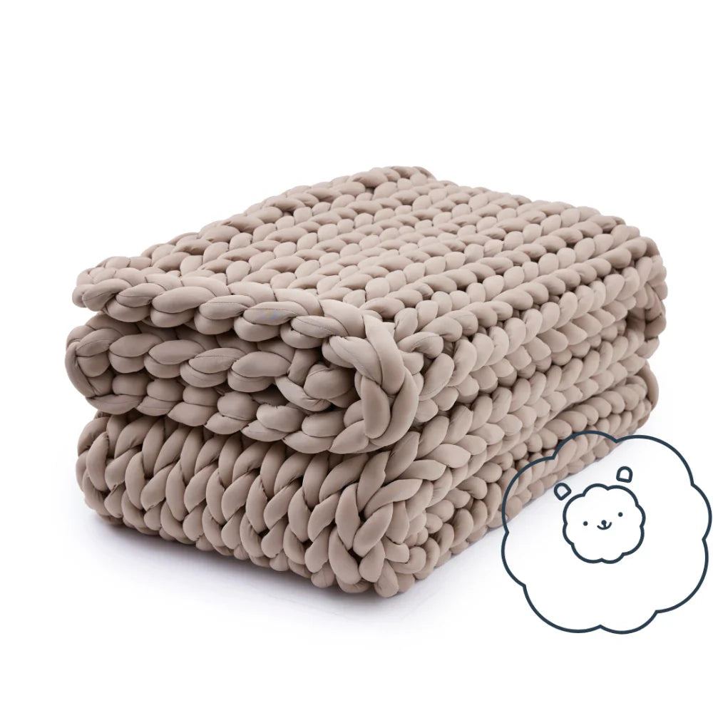 A chunky, hand-knit beige Ice Knit Weighted Blanket is neatly folded, with a cute cloud-like character in the corner hinting at its cozy comfort.