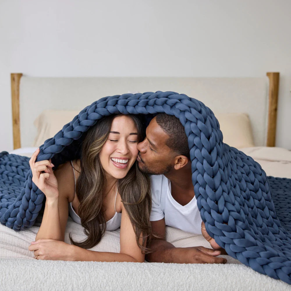 A smiling couple relaxes under the Ice Knit Weighted Blanket in bed; the man kisses the womans cheek as they appear cool, comfortable, and happy.