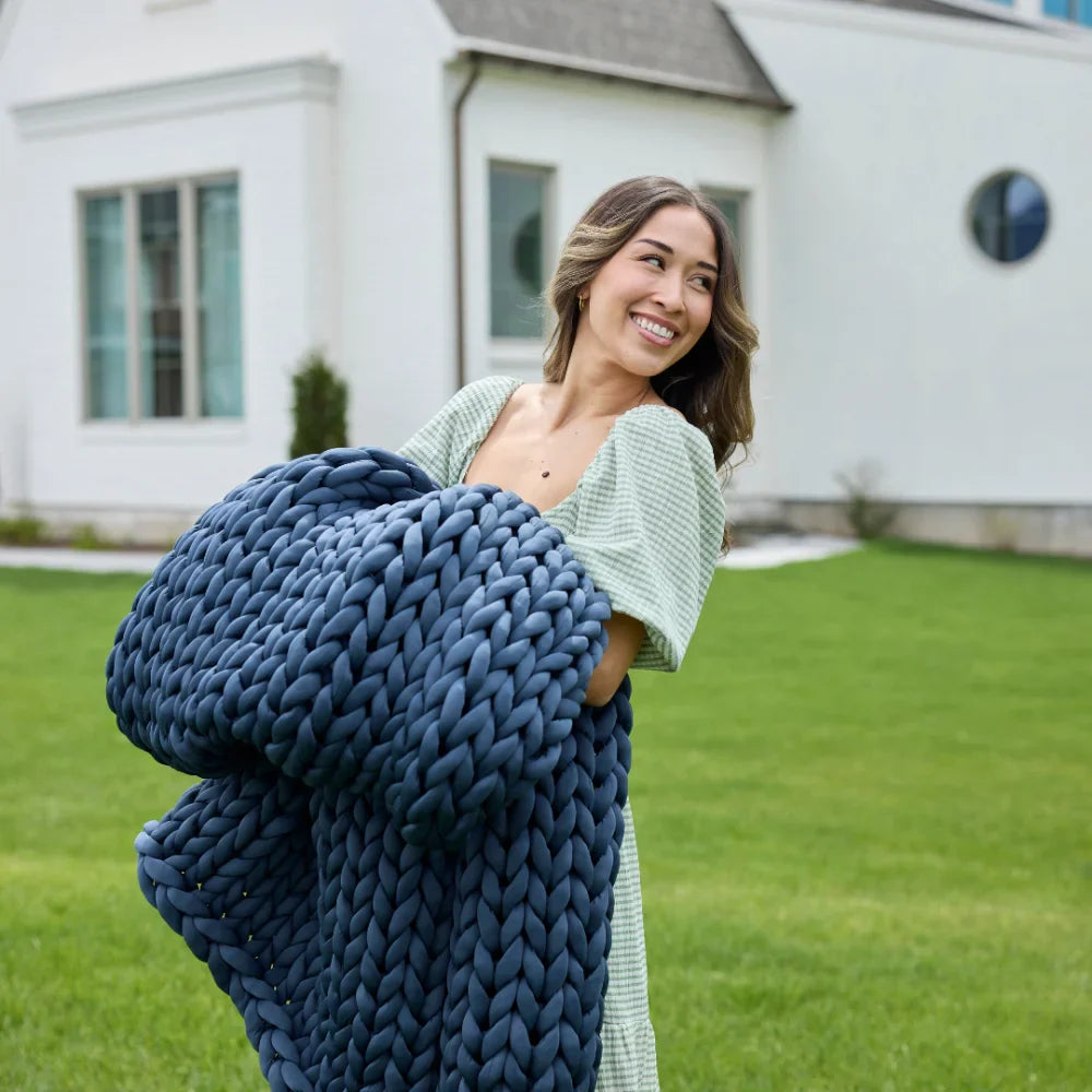 A woman stands on a lawn in front of a white house, smiling and holding the Ice Knit Weighted Blanket—a large, chunky blue blanket designed to keep you cool and comfortable.
