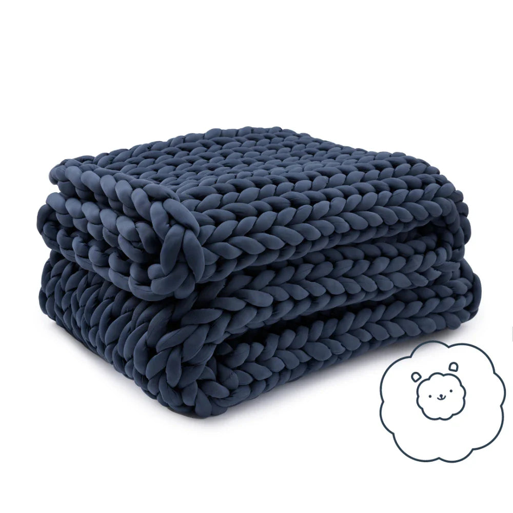 A neatly folded, chunky-knit Ice Knit Weighted Blanket in dark blue is shown. In the bottom right corner, theres a small illustration of a smiling sheep’s face inside a fluffy outline.