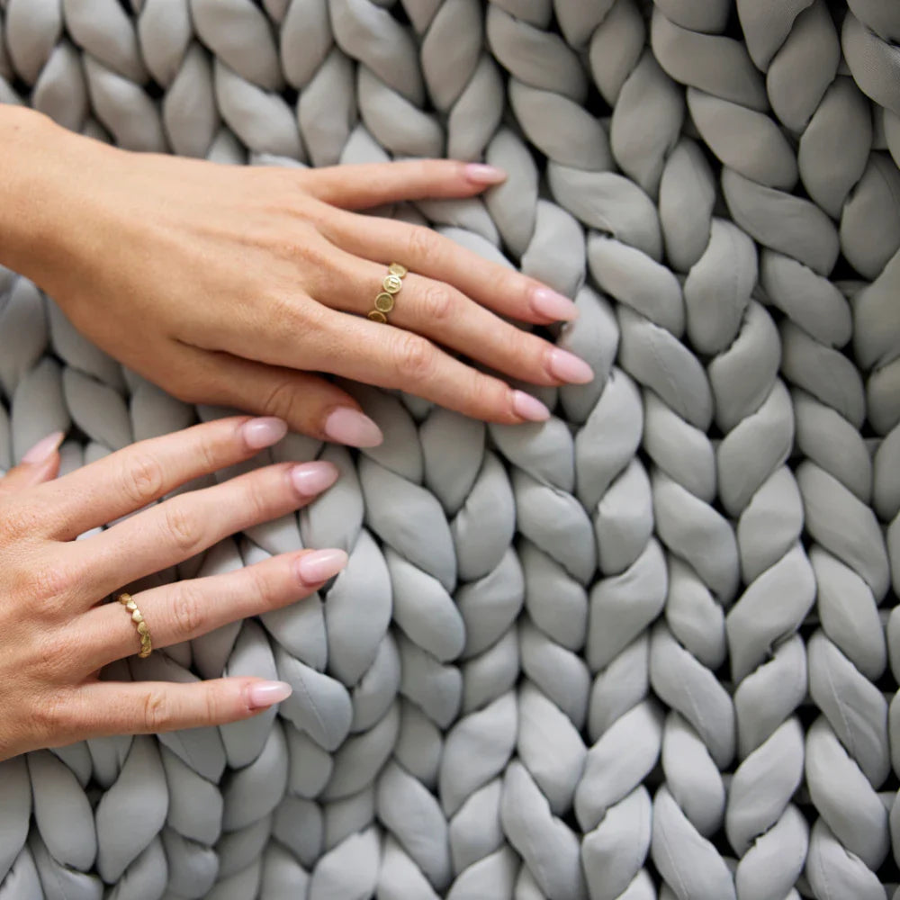Two hands with light pink manicured nails and gold rings rest on the Ice Knit Weighted Blanket in Silver Frost, featuring a thick, chunky-knit design.
