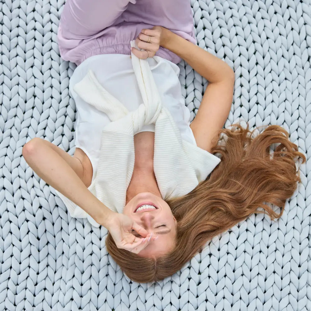 A smiling woman with long brown hair lies on the Ice Knit Weighted Blanket, wearing light purple pants and a white top with a sweater over her shoulders, making a peace sign over her eye.