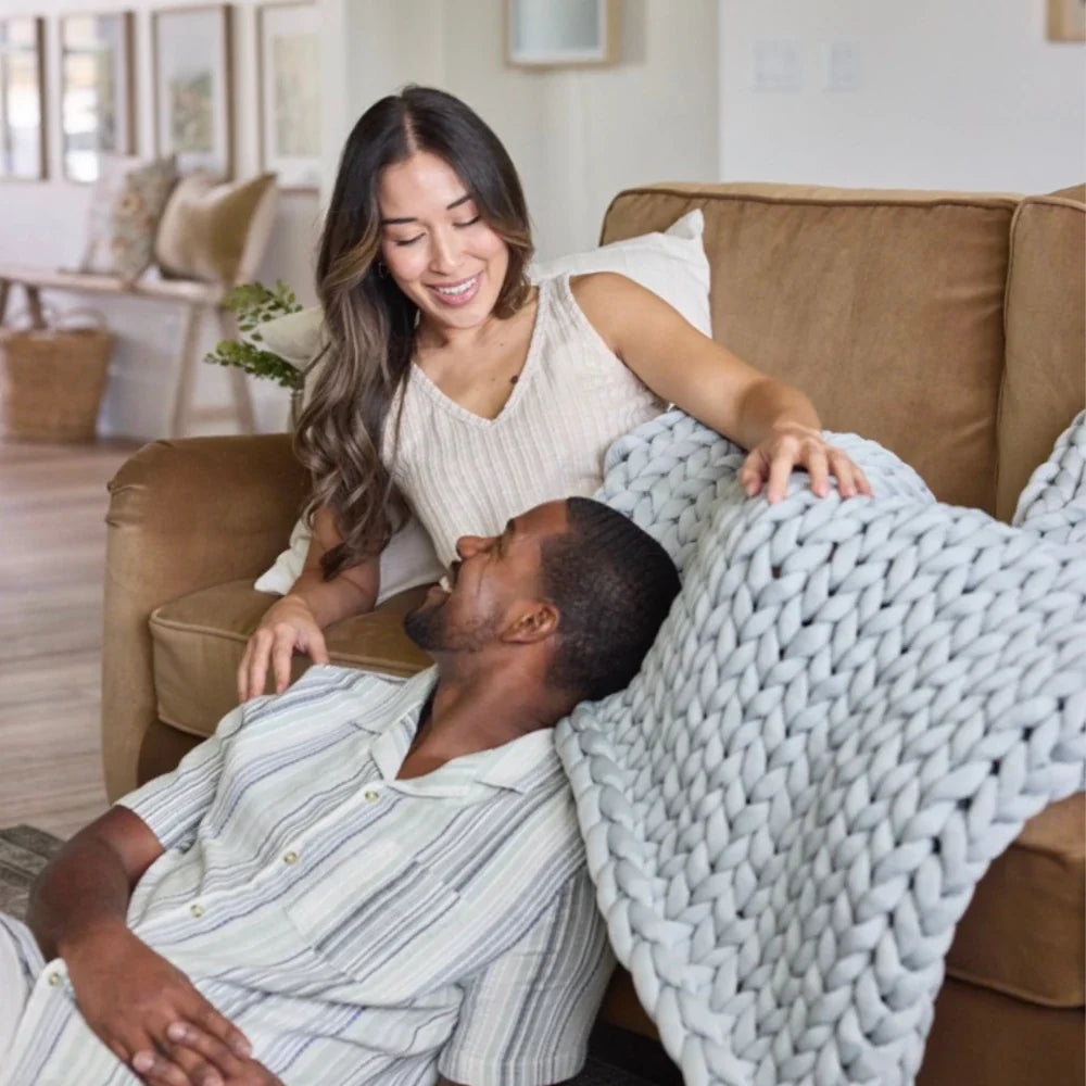 A smiling woman on the couch leans toward a man on the floor, both appearing relaxed and happy in a cozy living room, wrapped together in the Ice Knit Weighted Blanket amidst neutral decor.