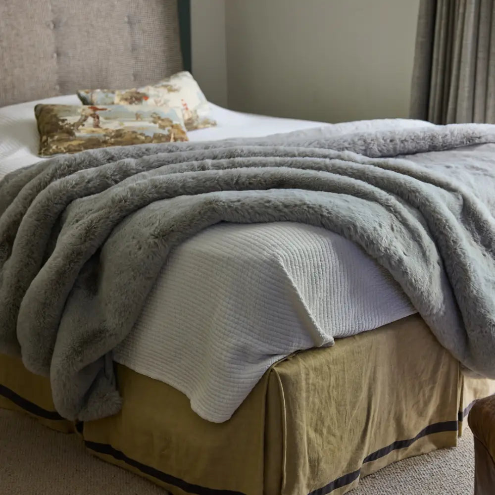 A softly lit bedroom features a cozy bed with a white Snuggle Luxe Blanket, a fluffy gray throw, a decorative pillow, and a beige bed skirt.
