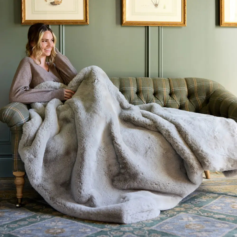 A woman smiles, wrapped in the Snuggle Luxe Blanket—a large, plush throw—while relaxing on a green plaid sofa in an elegant room decorated with framed art.