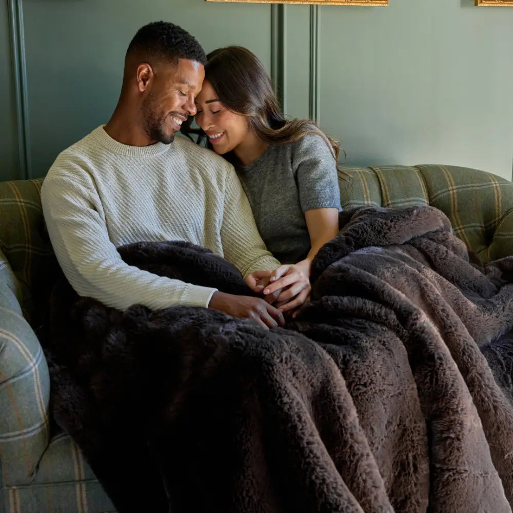 A couple smiles and holds hands on a couch, wrapped together in the cozy Snuggle Luxe Blanket.