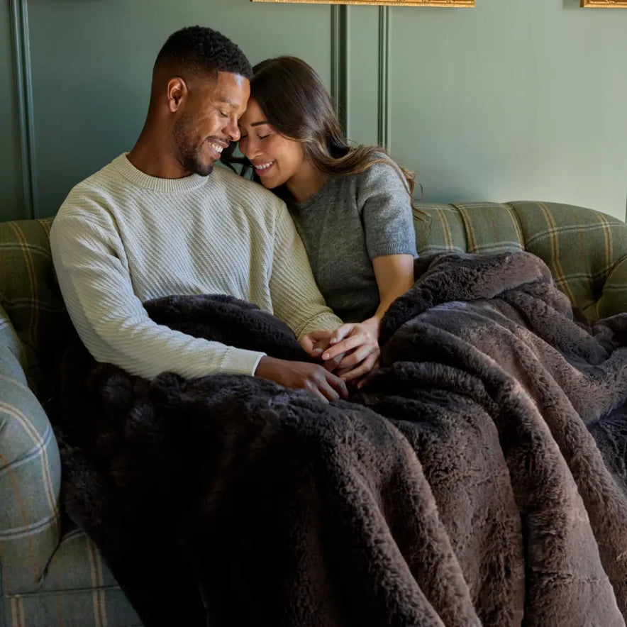 A couple smiles and holds hands on a couch, wrapped together in the cozy Snuggle Luxe Blanket.