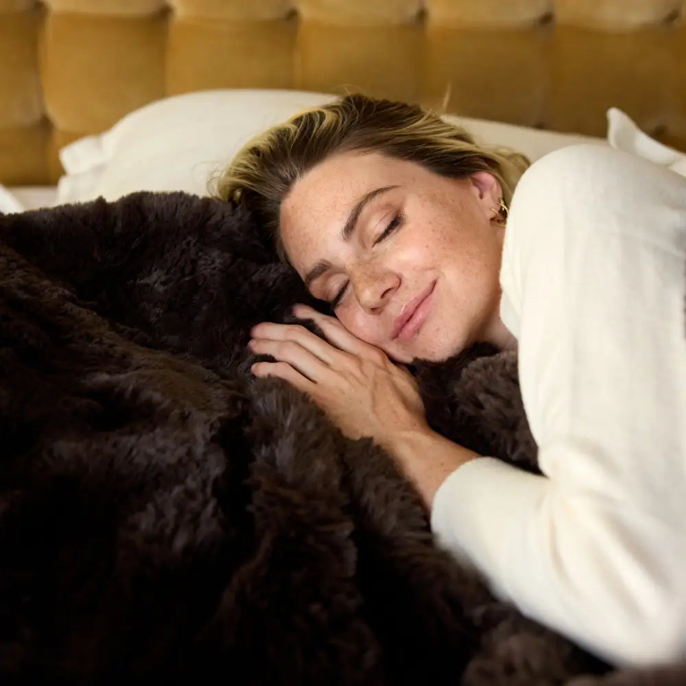 A person with closed eyes smiles while lying in bed, resting their head and hands on the Snuggle Luxe Blanket. Behind them is a tan tufted headboard and a white pillow.