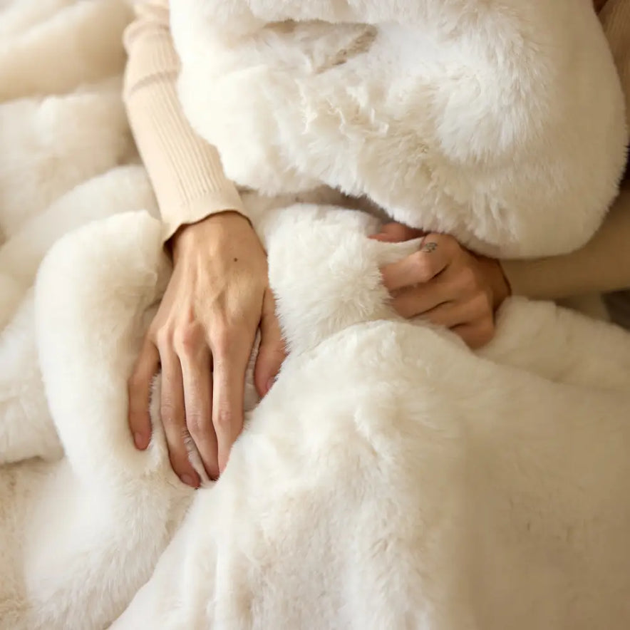 A person drapes themselves in the Snuggle Luxe Blanket, their hands resting on its soft, plush fabric, while wearing a beige sweater.