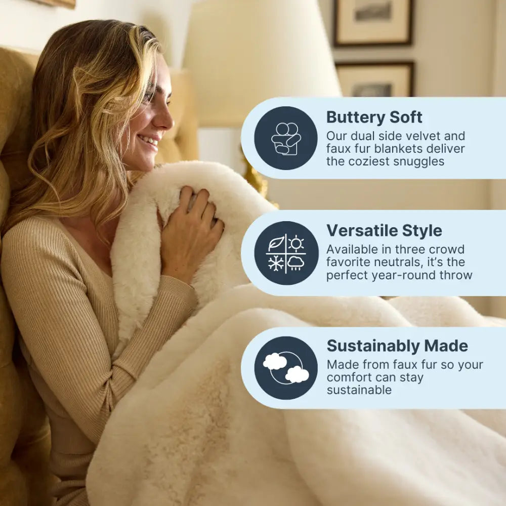 A woman smiles while wrapped in the Snuggle Luxe Blanket, a plush cream-colored faux fur throw thats buttery soft, sustainably made, and features versatile style—perfect for cozying up on the couch.
