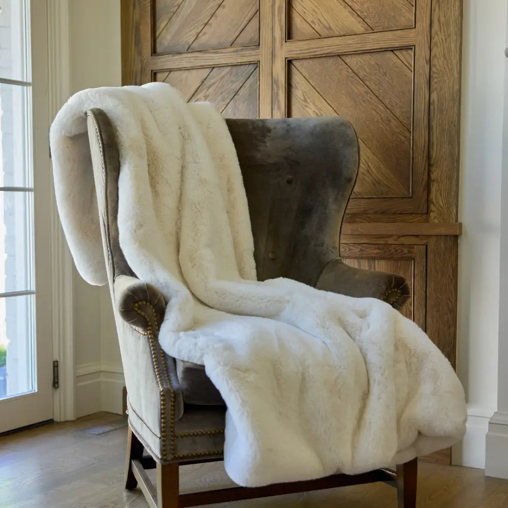 The Snuggle Luxe Blanket draped over a gray velvet armchair with brass studs, set on a wooden floor by a large window and wood-paneled cabinet, adds cozy elegance to the space.