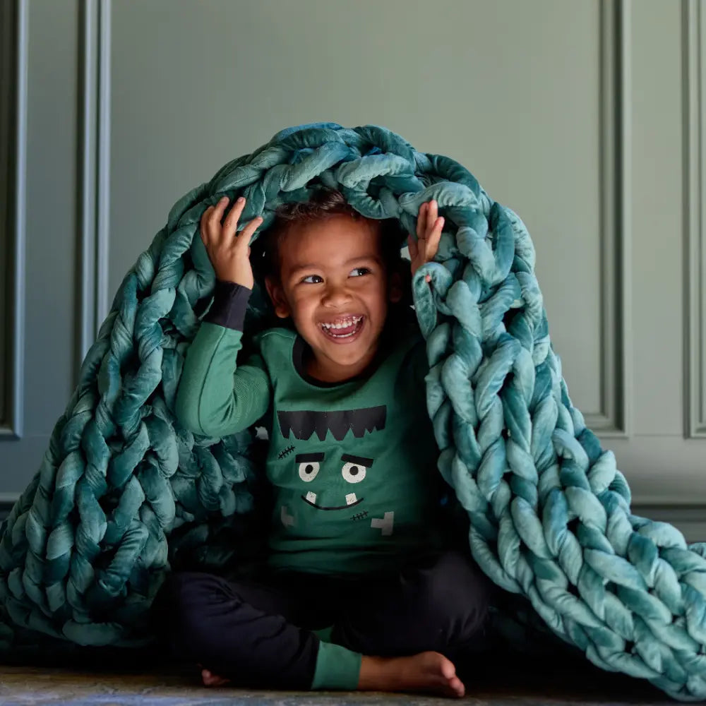 A smiling child in green cartoon pajamas sits with a Velvet Weighted Blanket draped over their head like a tent, adding cozy comfort against a light green paneled wall.