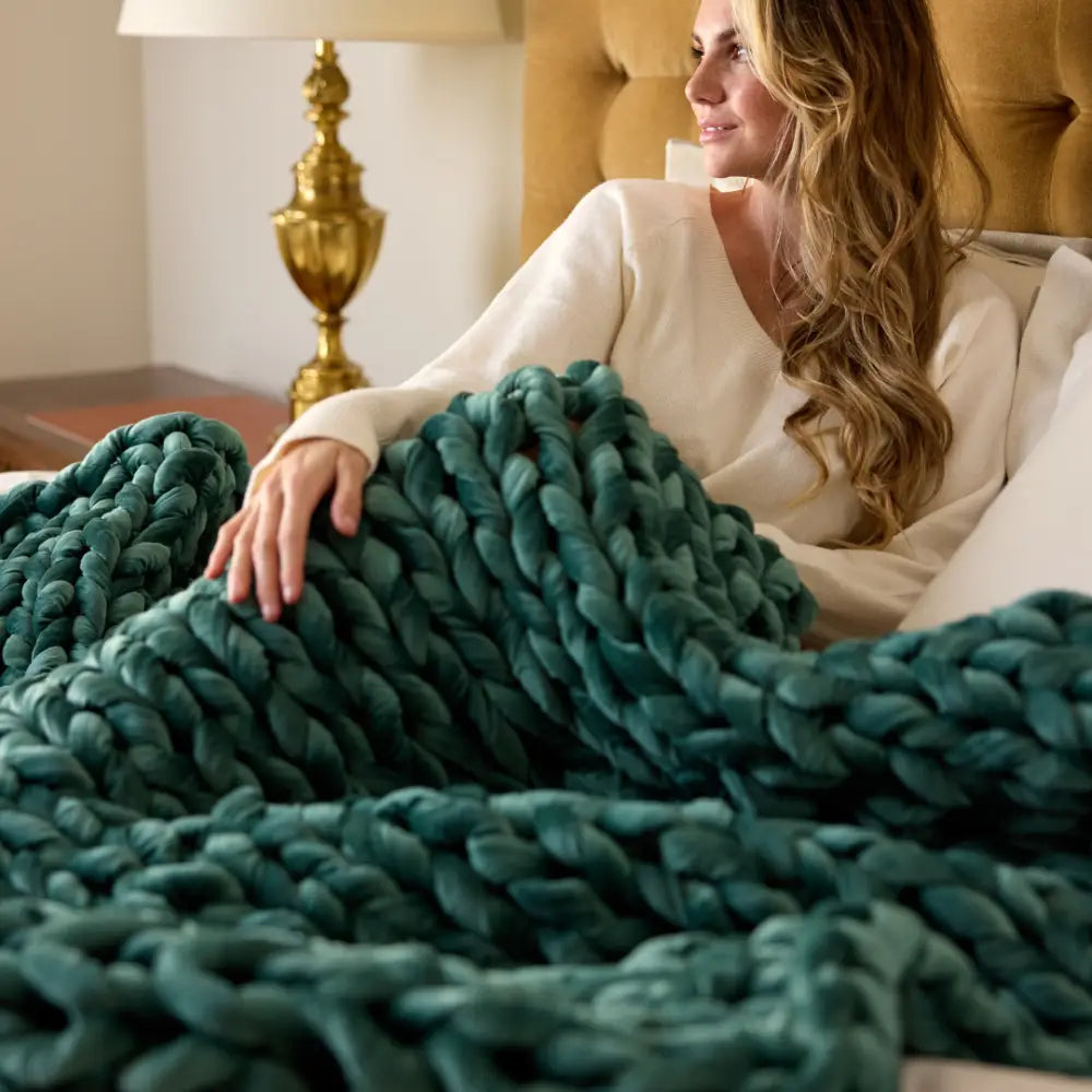 A woman sits in bed smiling, with a Velvet Weighted Blanket in chunky teal draped over her legs. A gold lamp and nightstand appear in the background.