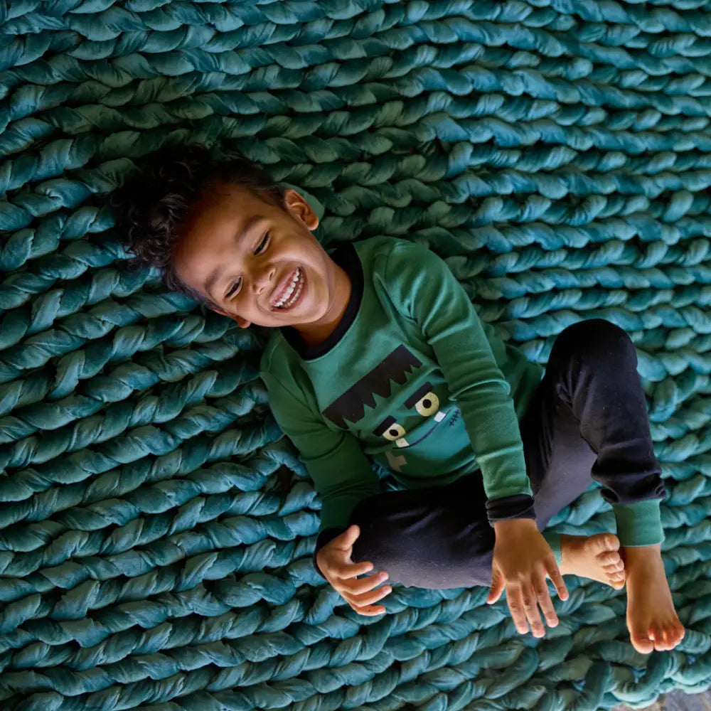 A young child in green and black pajamas laughs while lying on a teal knit rug, wrapped in cozy comfort under the Velvet Weighted Blanket, which features ultra-soft velvet for extra warmth.