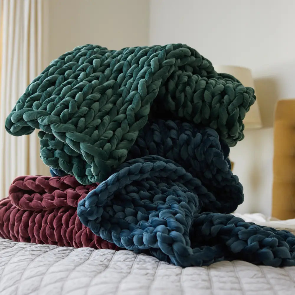 A velvet weighted blanket in green, navy blue, and burgundy hues is stacked on a bed with a white quilted cover for cozy comfort. A blurred lamp and headboard appear in the background.