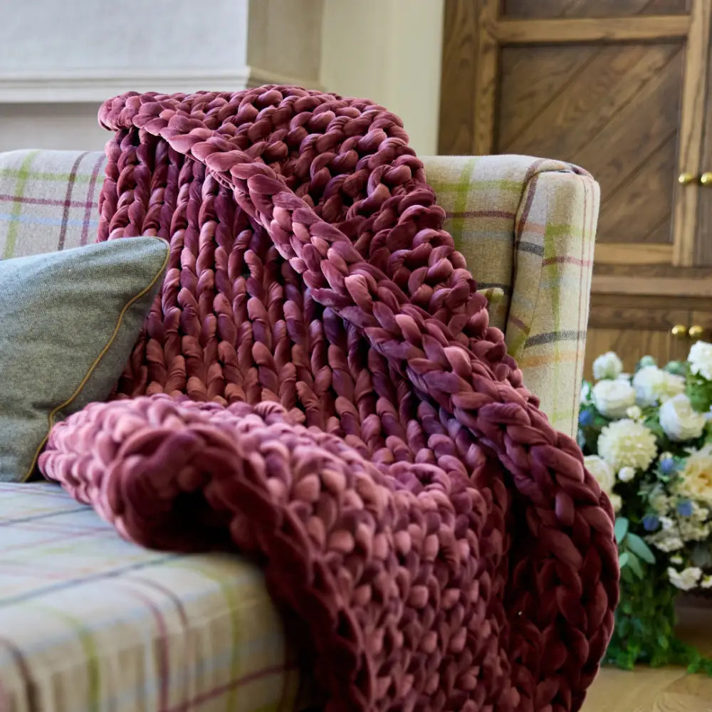 A plush Velvet Weighted Blanket in burgundy adds cozy comfort draped over a plaid sofa, paired with a gray cushion and a bouquet of white and blue flowers in the background.