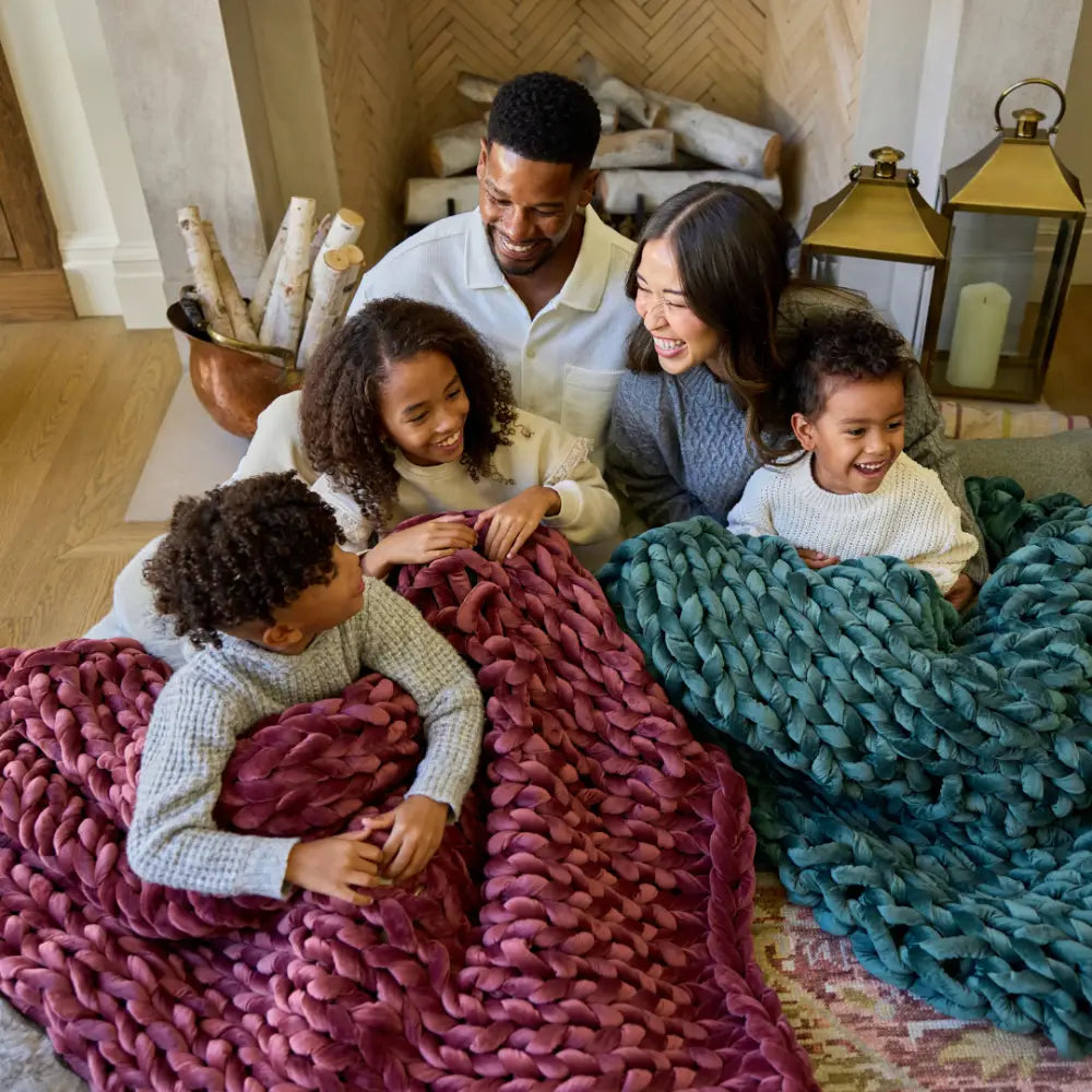A smiling family of four sits on the floor in front of a fireplace, wrapped in cozy Velvet Weighted Blankets—one maroon, one teal—enjoying comfort, laughter, and time together.