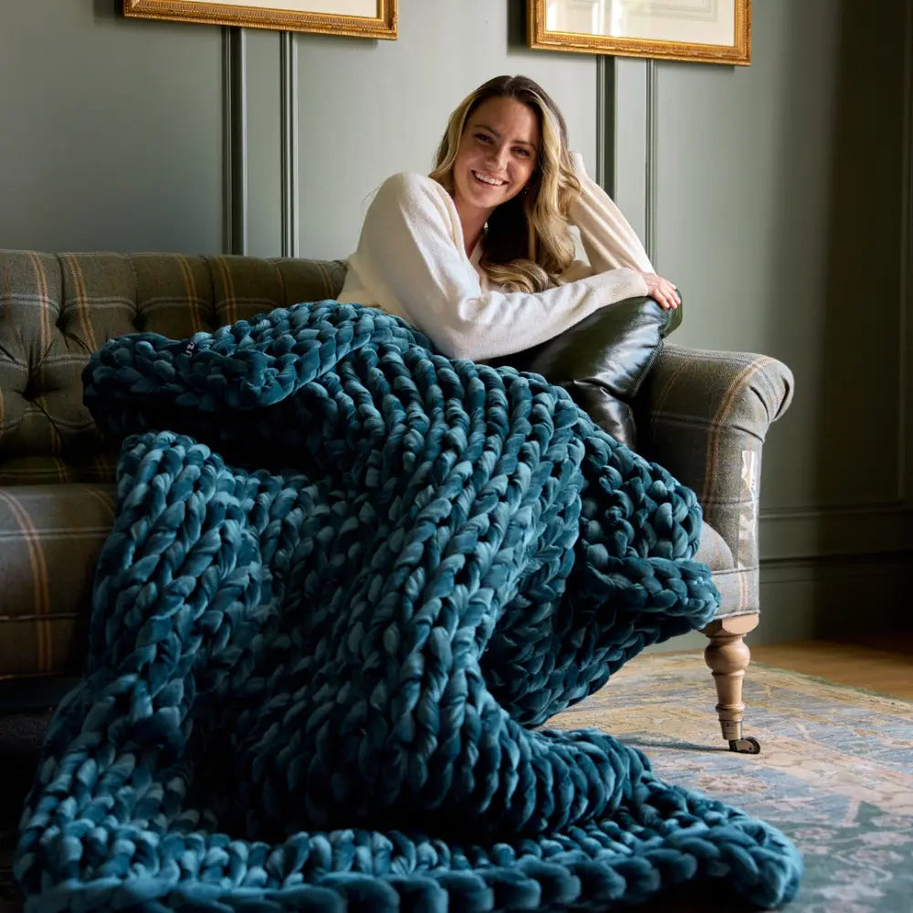 A woman with long hair sits on a sofa, smiling, wrapped in a sapphire Velvet Weighted Blanket. Framed pictures on the wall and a patterned rug enhance the cozy comfort of the room.