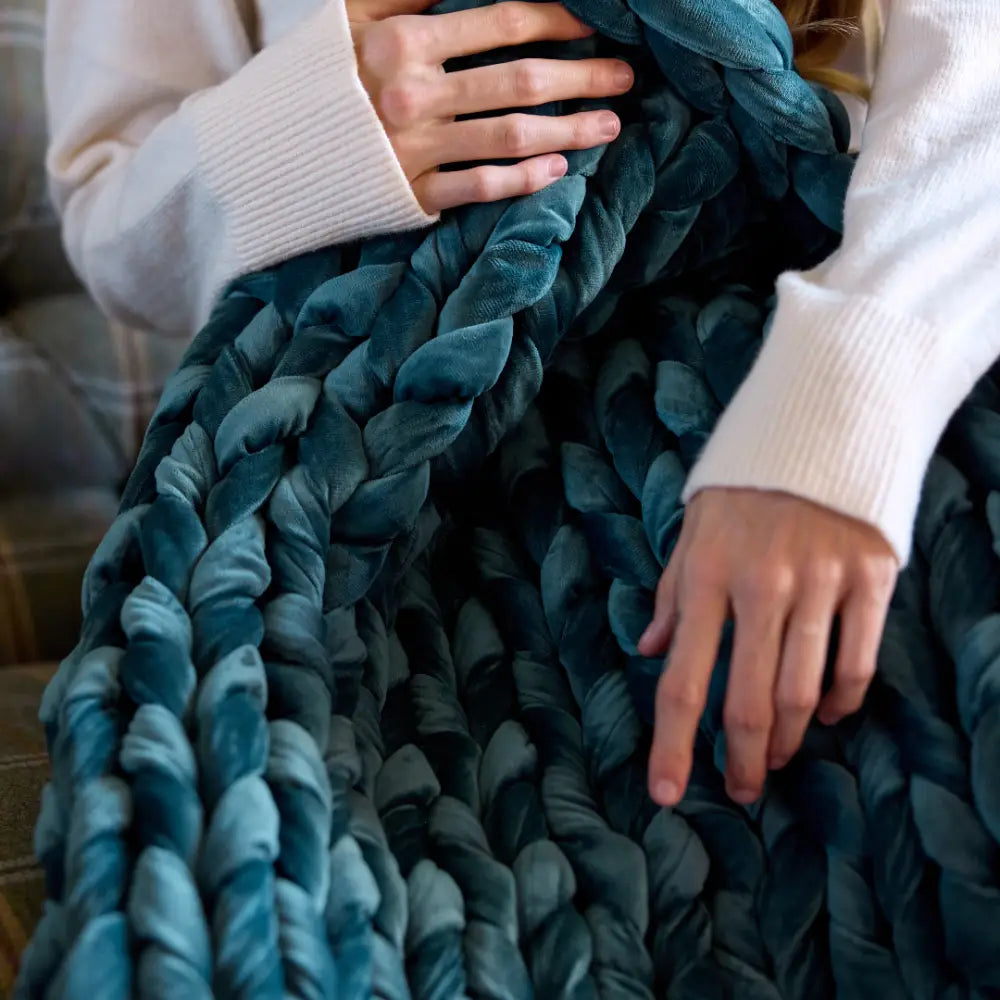 Close-up of a person’s hands holding the Velvet Weighted Blanket in teal. They wear a cream sweater and sit on a plaid couch, enjoying the ultra-soft, chunky comfort.