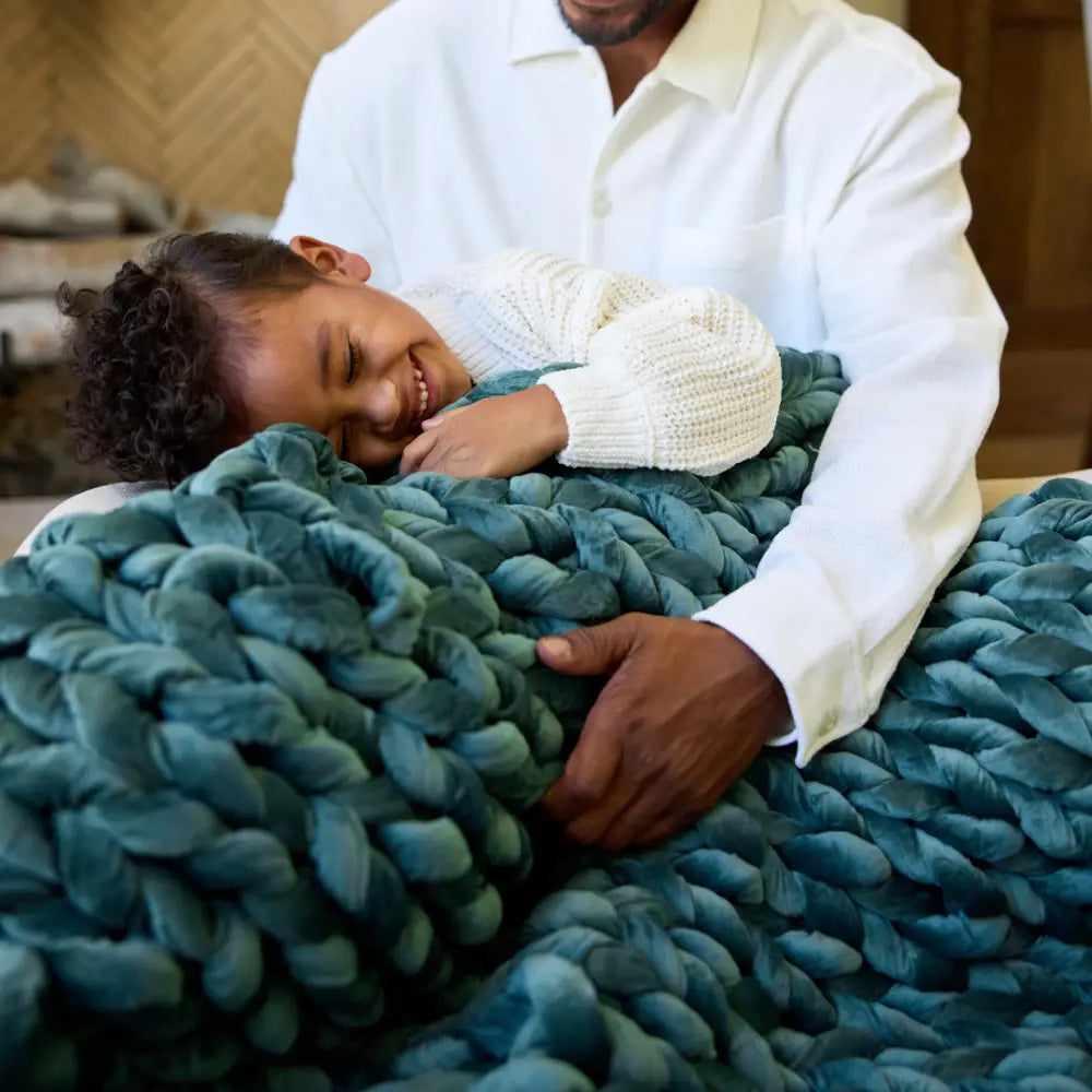 A young child smiles and cuddles under the Velvet Weighted Blanket while being embraced by an adult in a white shirt, creating a cozy, affectionate scene filled with warmth and comfort.
