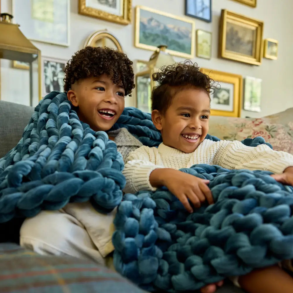 Two smiling children sit on a couch, wrapped together in a Velvet Weighted Blanket. Framed artwork decorates the wall behind them, creating a cozy and cheerful atmosphere.
