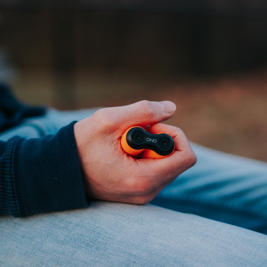 A person in a dark blue sleeve holds a small orange and black ONO Roller Soft Touch Plastic fidget toy for stress relief while sitting outdoors on light blue jeans, with the background blurred.