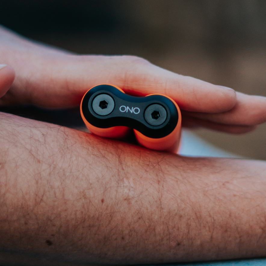 A hand holds an orange ONO Roller Soft Touch Plastic fidget toy against a forearm, parallel to the arm. The device has black ends marked ONO and is perfect for stress relief.