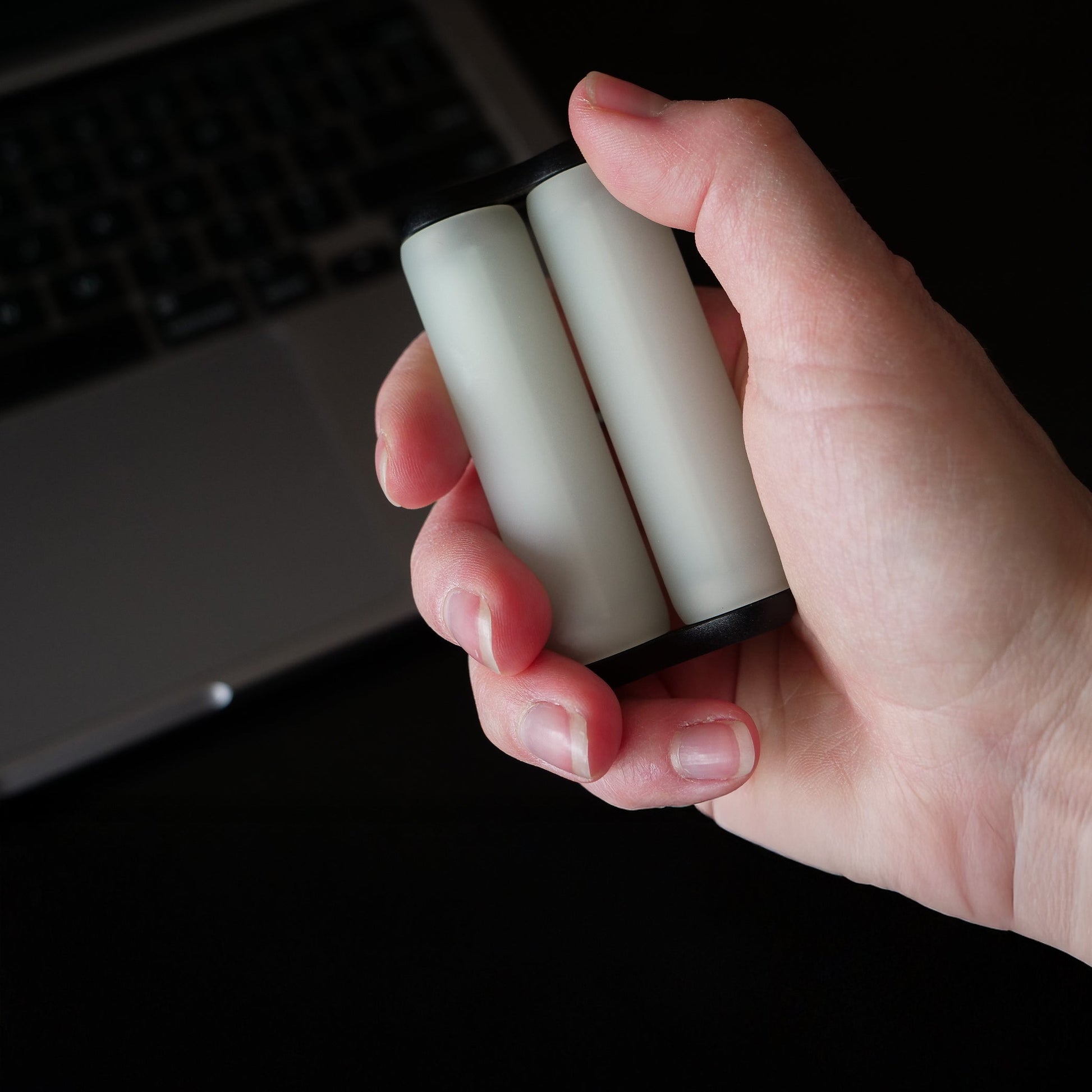 A hand holds the ONO Roller Soft Touch Plastic, a small white and black cylindrical fidget toy with two silent rollers, in front of a laptop keyboard—ideal for stress relief during work.