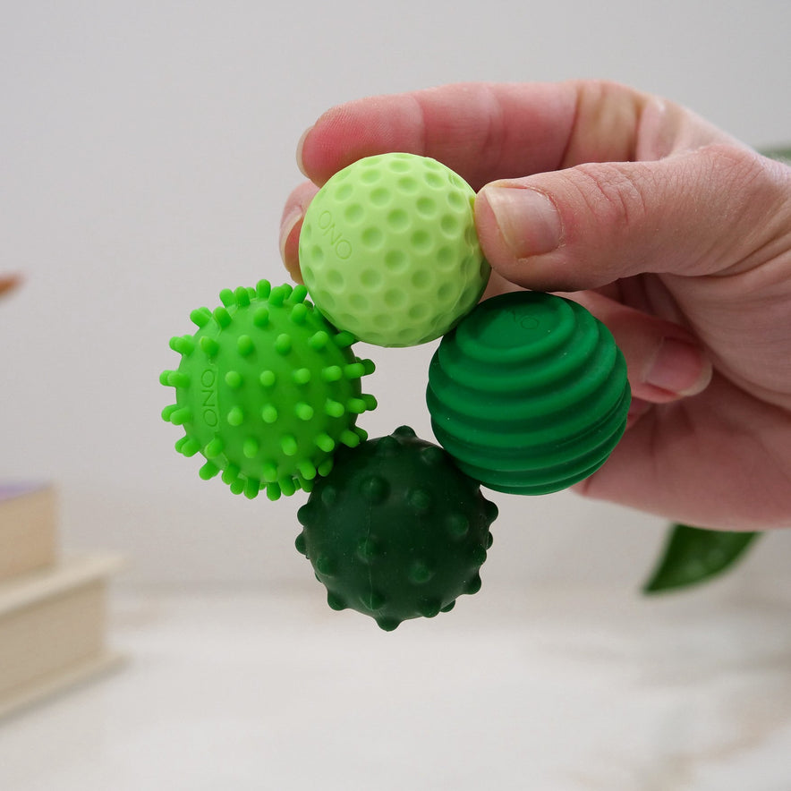A hand holds four green Signets—small textured fidget balls featuring bumps, spikes, ridges, and round nubs. The background is neutral with a plant and books in view.