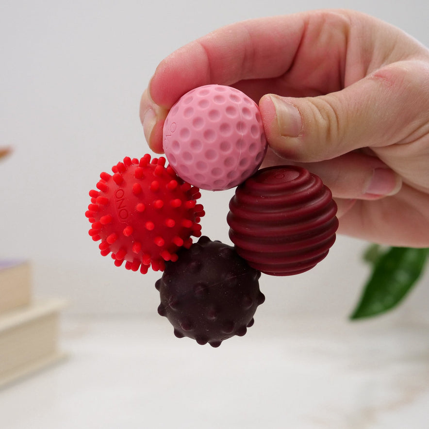 A hand holds four Signets balls, each offering a unique fidgeting experience: pink and dimpled, red and spiky, dark brown and bumpy, and maroon with ridges—perfect for hands-on play that aids focus.