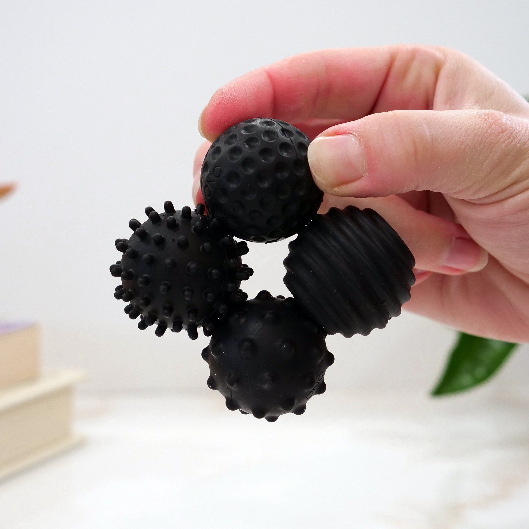 A hand holds a black Signets fidget toy: four connected silicone-coated magnetic balls, each with a unique textured surface—bumps, spikes, ridges, and dots. A blurred book and green plant are seen in the background.