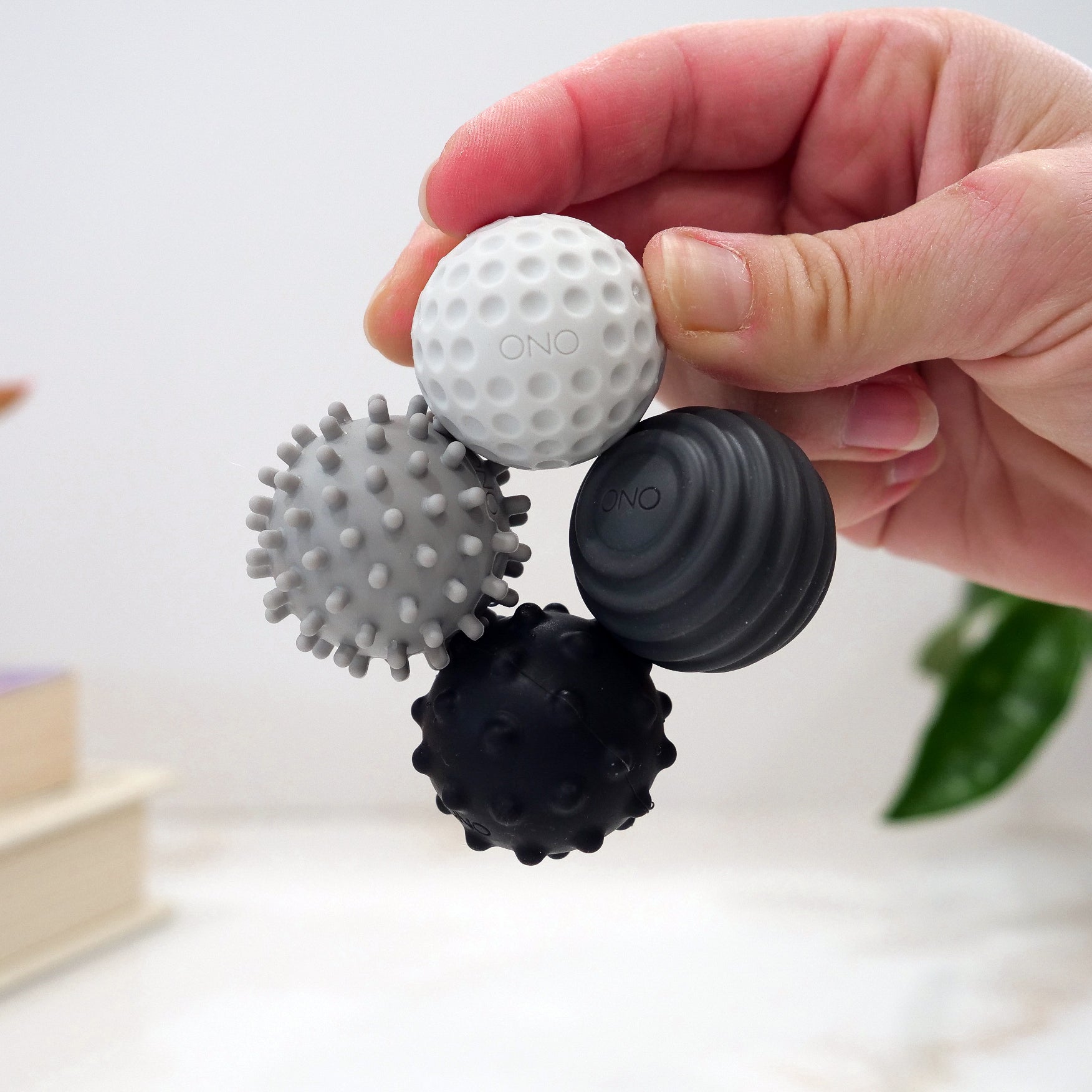 A hand holds Signets, a fidget toy featuring four connected, silicone-coated magnets in varying textures and shades from white to black. A blurred book and green plant in the background enhance the fidgeting experience.