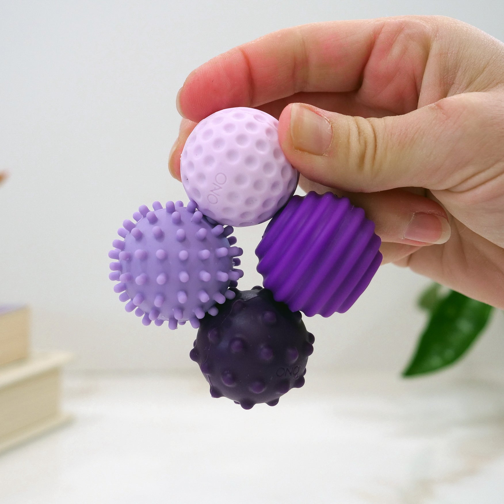 A hand holds four Signets—small, textured sensory balls in various purple shades with bumpy and ridged surfaces—designed to enhance fidgeting and focus. A blurred plant and books appear in the background.