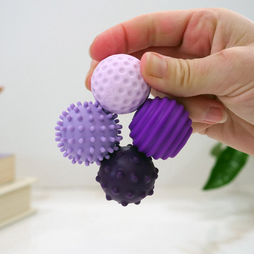 A hand holds four Signets—small, textured sensory balls in various purple shades with bumpy and ridged surfaces—designed to enhance fidgeting and focus. A blurred plant and books appear in the background.