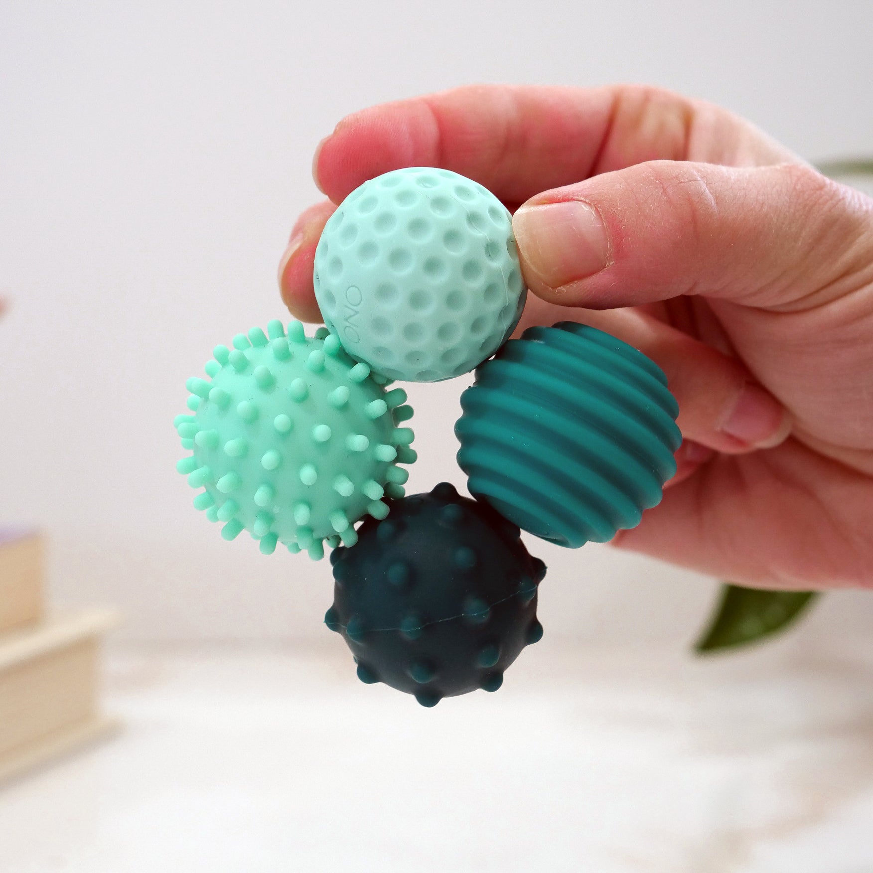 A hand holds four Signets—small sensory balls in blue and green, each with unique bumpy, spiky, ridged, or dimpled textures—for a calming fidgeting experience that aids focus. The background is softly blurred.